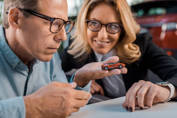 Close-up shot of adult man and smiling female car dealer looking at toy car in showroom — Stock Photo