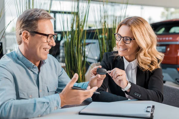 Happy adult man and female car dealer passing car key in showroom — Stock Photo