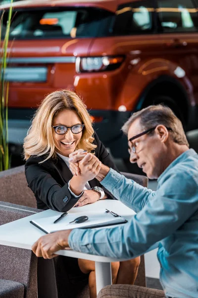 Adult man and female car dealer arm wrestling in showroom — Stock Photo