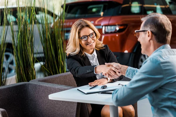 Happy adult customer and female car dealer shaking hands in showroom — Stock Photo