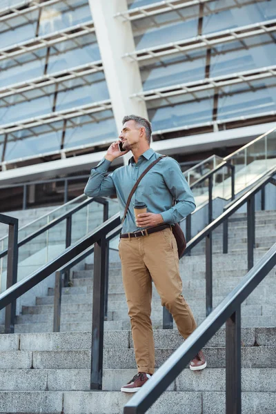 Красивий чоловік розмовляє по смартфону і дивиться геть, стоячи з кавою, щоб йти по сходах — стокове фото