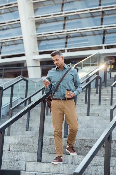 Handsome middle aged man holding paper cup and using smartphone on stairs — Stock Photo