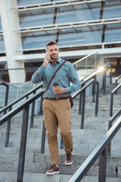 Bonito meia idade empresário segurando café para ir e falando por smartphone em escadas — Fotografia de Stock