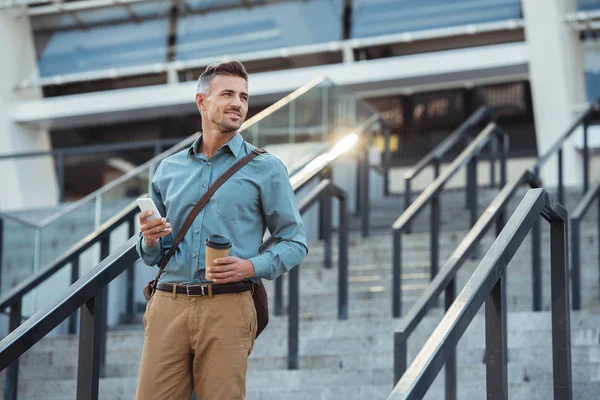 Homme d'affaires d'âge moyen souriant avec café à emporter et smartphone regardant loin dans les escaliers — Photo de stock
