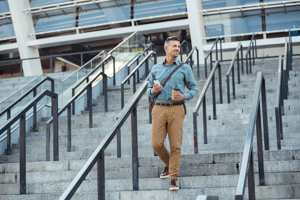 Smiling middle aged businessman holding paper cup and smartphone while walking down on stairs — Stock Photo