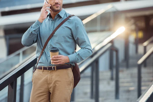 Cropped shot of businessman holding coffee to go and talking by smartphone on stairs — Stock Photo