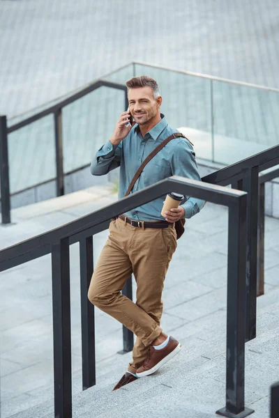 Високий кут зору усміхненого чоловіка, який тримає каву, щоб піти і розмовляти по смартфонах на сходах — Stock Photo