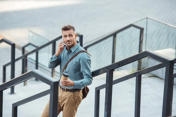 Sourire homme d'âge moyen parlant par smartphone main tenant tasse en papier tout en se tenant debout sur les escaliers — Photo de stock