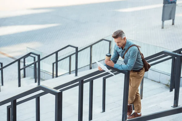Blick aus der Vogelperspektive auf einen Geschäftsmann mittleren Alters, der einen Coffee to go hält und Zeitung liest, während er sich an Geländer auf der Treppe lehnt — Stockfoto
