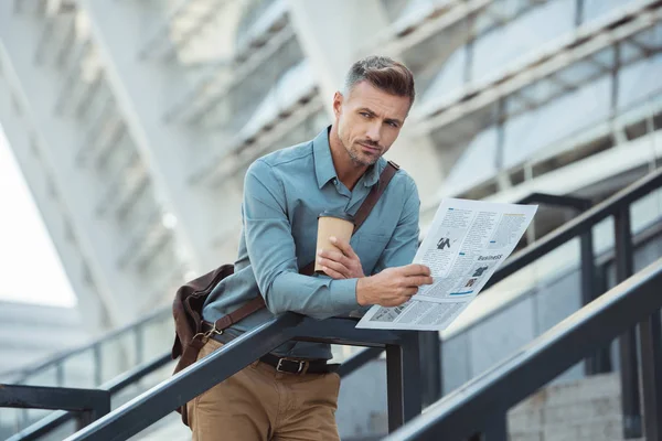 Apuesto hombre de mediana edad sosteniendo café para ir y periódico mientras está de pie en las escaleras - foto de stock