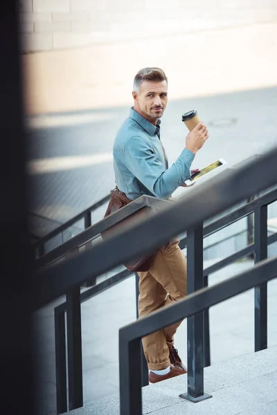 Избирательный фокус привлекательного бизнесмена средних лет, пьющего кофе из бумажной чашки и использующего цифровые планшеты на улице — стоковое фото