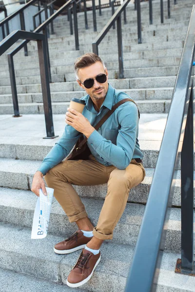 Усміхнений чоловік в сонцезахисних окулярах тримає газету і п'є каву з паперової чашки на сходах — стокове фото
