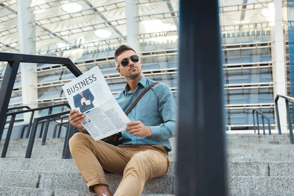 Bell'uomo in occhiali da sole che tiene il giornale aziendale e distoglie lo sguardo mentre si siede sulle scale — Foto stock