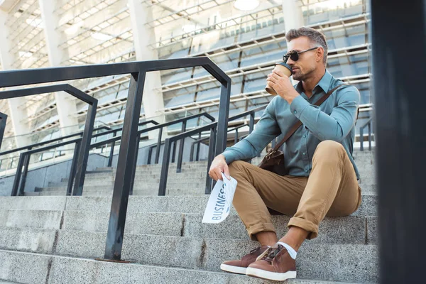 Bel homme dans les lunettes de soleil tenant journal d'affaires et boire du café de tasse en papier tout en étant assis sur les escaliers — Photo de stock