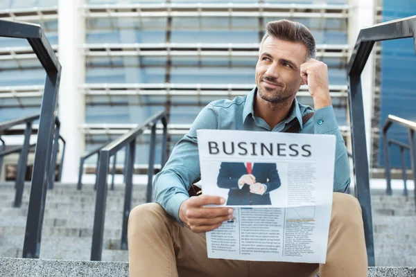 Pensive smiling man holding business newspaper and looking away while sitting on stairs — Stock Photo