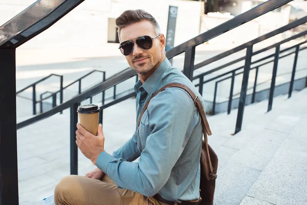 Красивый улыбающийся мужчина средних лет в солнцезащитных очках сидит на лестнице и держит кофе, чтобы пойти — стоковое фото