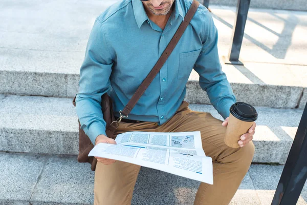 Cropped shot of businessman with coffee to go sitting on stairs and reading newspaper — Stock Photo