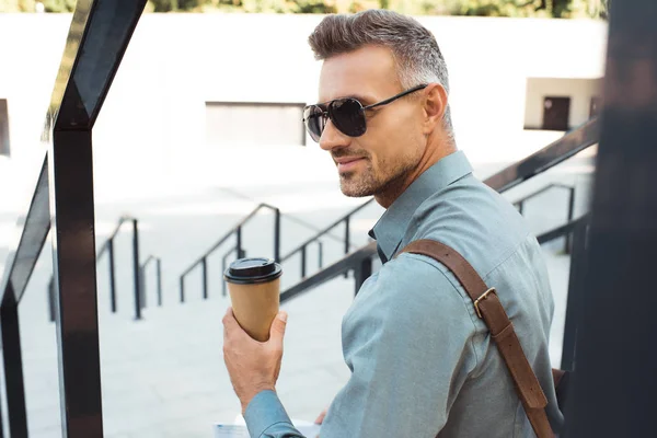 Bel homme souriant dans des lunettes de soleil tenant du café pour aller et assis sur les escaliers — Photo de stock