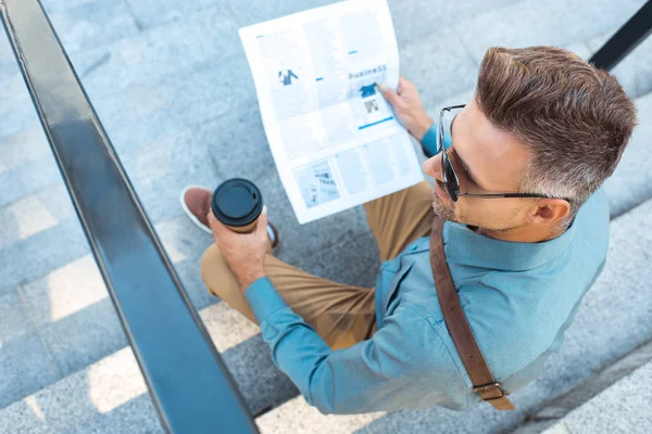 Vue grand angle de l'homme en lunettes de soleil tenant tasse en papier tout en étant assis sur les escaliers et en lisant le journal — Photo de stock