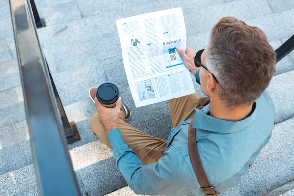 High angle view of man with paper cup sitting on stairs and reading newspaper — Stock Photo