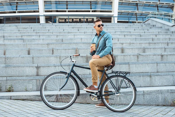 Красивый мужчина в солнечных очках сидит на велосипеде с кофе, чтобы пойти и оглянуться через плечо на улице — стоковое фото