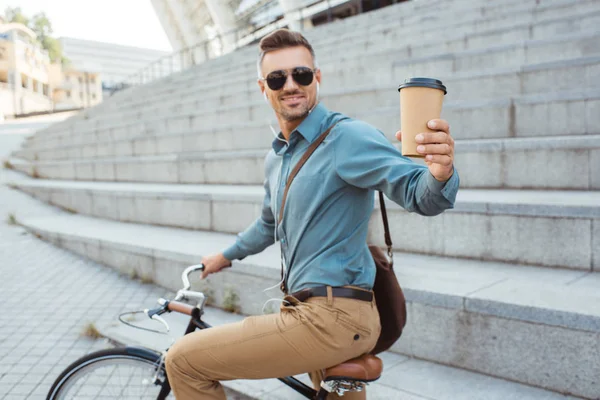 Красивый улыбчивый мужчина, держащий кофе, чтобы покататься на велосипеде по улице — стоковое фото