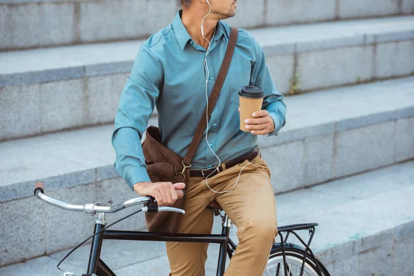 Cropped shot of man in earphones holding paper cup and sitting on bicycle — Stock Photo