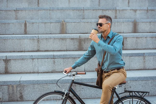 Красивый мужчина в солнечных очках на велосипеде и пьет из бумажной чашки на улице — стоковое фото