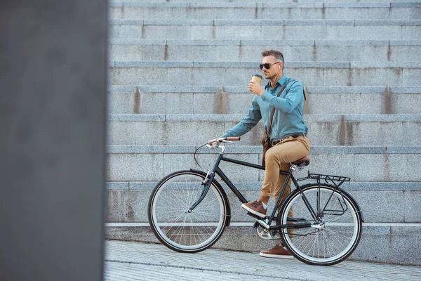 Handsome man riding bicycle and drinking from paper cup on street — Stock Photo