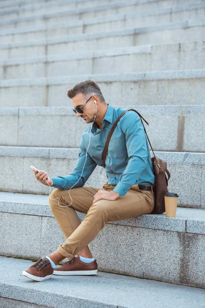 Handsome middle aged man in sunglasses and earphones sitting on stairs and using smartphone — Stock Photo