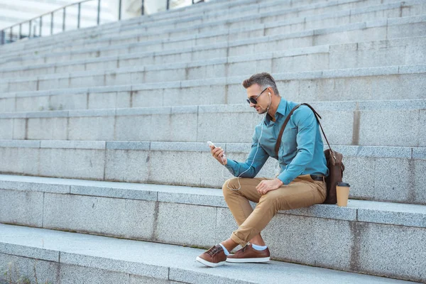 Schöner, stylischer Mann mit Kopfhörer und Sonnenbrille, der auf der Treppe sitzt und das Smartphone benutzt — Stockfoto