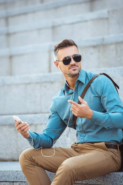 Handsome middle aged man in earphones sitting on stairs and using smartphone — Stock Photo