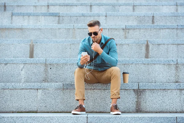 Stylish middle aged man in earphones sitting on stairs and using smartphone — Stock Photo