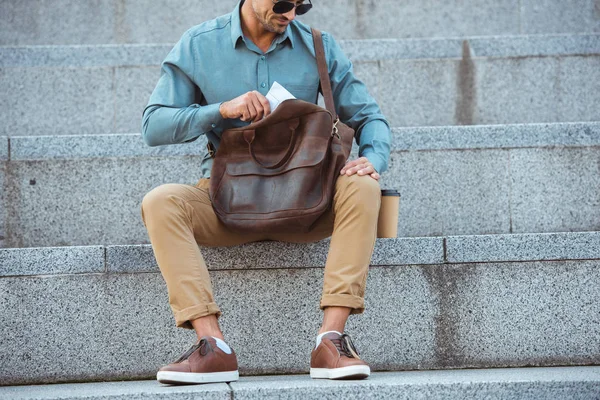 Cropped shot of man in sunglasses sitting on stairs and putting newspaper into leather bag — Stock Photo