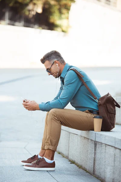 Vista lateral del hombre en auriculares y gafas de sol sentado en las escaleras y utilizando el teléfono inteligente - foto de stock