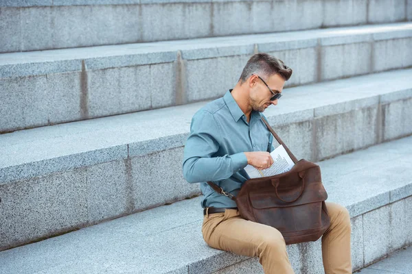 Bel homme élégant d'âge moyen assis sur les escaliers et mettre du journal dans un sac en cuir — Photo de stock