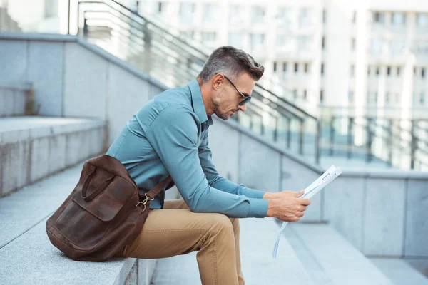 Side view of stylish middle aged businessman sitting in stairs and reading newspaper — Stock Photo