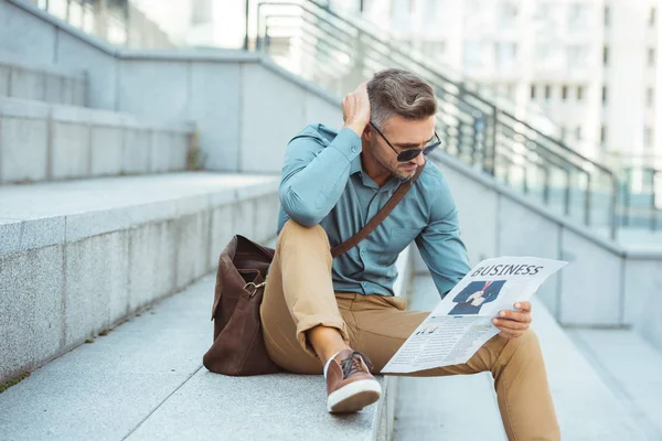 Hombre de mediana edad elegante sentado en las escaleras y leyendo el periódico de negocios - foto de stock