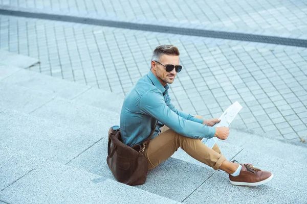 Handsome stylish man in sunglasses reading newspaper on stairs — Stock Photo