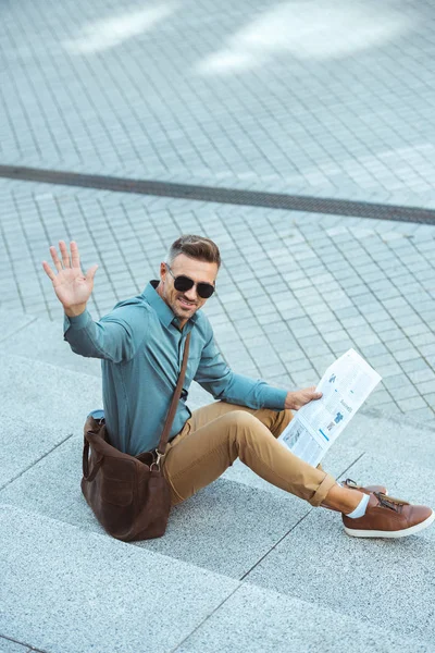 Sorrindo homem de meia idade sentado em escadas com jornal e acenando mão — Fotografia de Stock