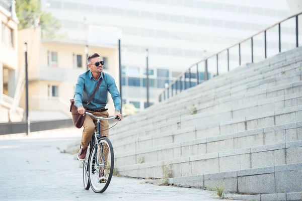 Guapo hombre de negocios de mediana edad en gafas de sol montar en bicicleta y mirando hacia la calle - foto de stock