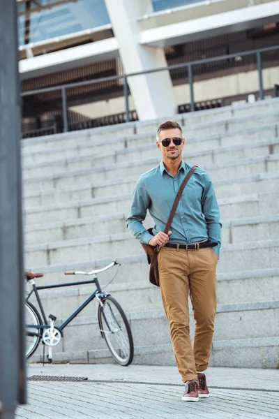 Красивый мужчина средних лет в солнечных очках, улыбающийся в камеру во время прогулки по улице, велосипед позади — стоковое фото