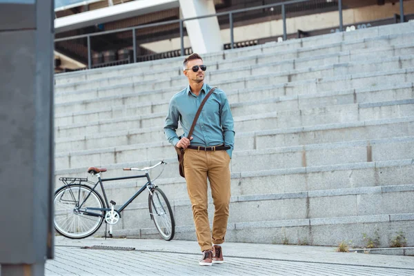 Handsome middle aged businessman in sunglasses walking on street, bicycle on background — Stock Photo