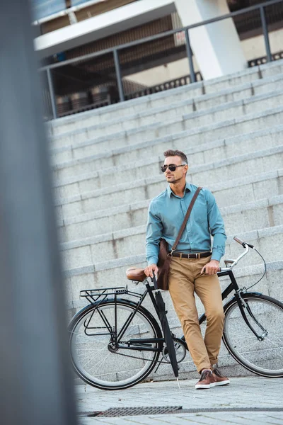 Handsome middle aged businessman in sunglasses holding umbrella while standing near bicycle on street — Stock Photo