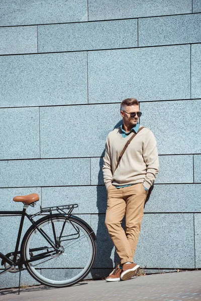 Elegante hombre de mediana edad en gafas de sol de pie con las manos en los bolsillos cerca de la bicicleta en la calle - foto de stock