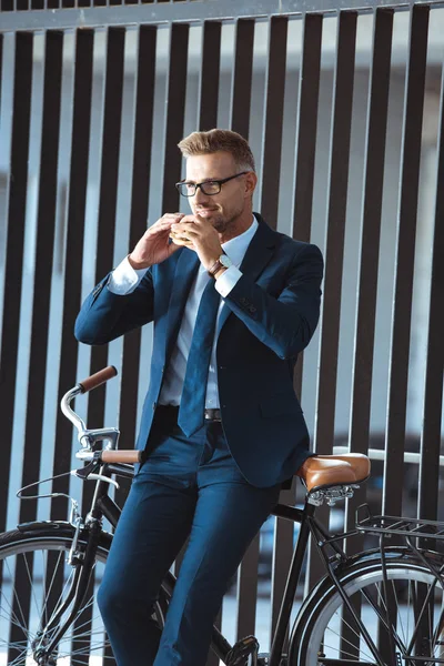 Усміхнений красивий бізнесмен середнього віку в костюмі сидить на велосипеді і їсть бургер на вулиці — стокове фото