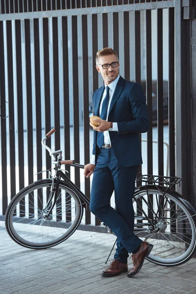 Smiling middle aged businessman leaning at bicycle and holding hamburger on street — Stock Photo