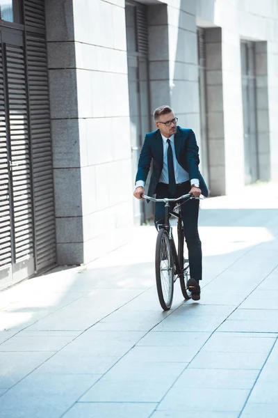 Handsome middle aged businessman in suit and eyeglasses riding bicycle on street — Stock Photo