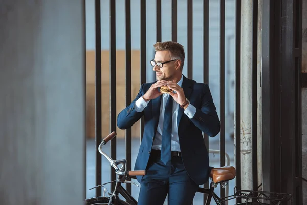 Smiling middle aged businessman in eyeglasses sitting on bike and holding hamburger — Stock Photo