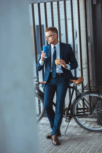 Ganzkörperansicht des gutaussehenden Geschäftsmannes, der Hamburger hält und aus Pappbechern trinkt, während er auf dem Fahrrad sitzt und wegschaut — Stockfoto